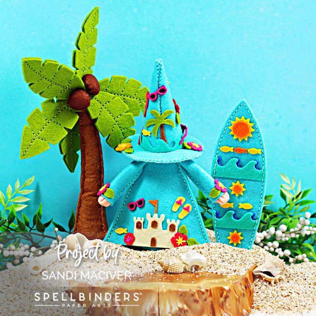 Back view of a handmade tropical Fergus felt gnome standing on a sandy beach beside a palm tree and surfboard. The aqua gnome features a detailed sandcastle scene on his cloak, along with beach accessories, sunglasses, seashells, tropical flowers, and summer-themed embellishments. The coordinating surfboard is decorated with sunshine, waves, and colorful fish for a fun coastal display.