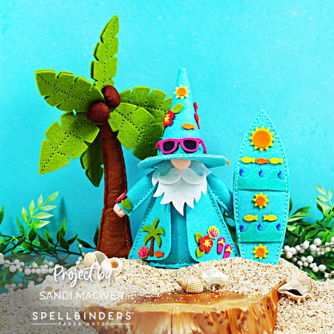 Tropical Fergus felt gnome decor standing on a sandy beach beside a palm tree and surfboard. The aqua-colored gnome wears bright pink sunglasses and a flower-adorned hat, while the matching surfboard features sunny beach-themed details. Seashells and greenery complete this cheerful summer felt decoration.