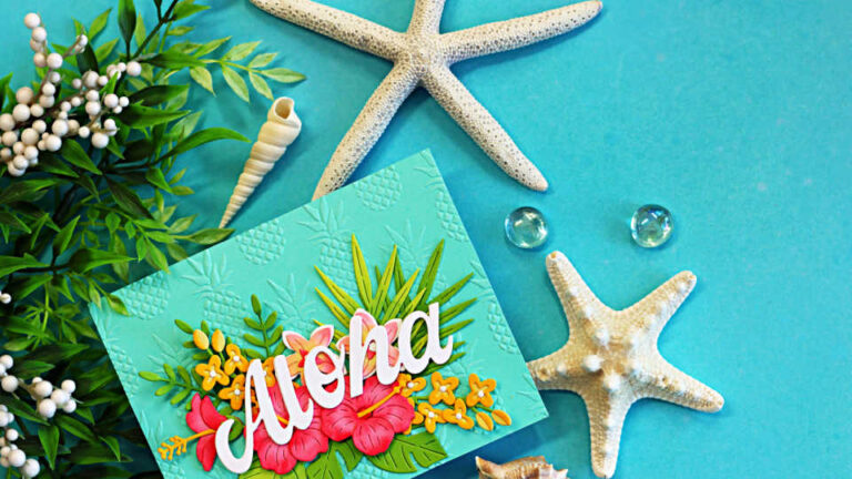 Handmade tropical Aloha card featuring layered hibiscus flowers, tropical leaves, and a large white “Aloha” sentiment on an aqua embossed background. The card is styled with starfish, seashells, greenery, and glass gems on a bright turquoise backdrop, creating a relaxing island-inspired scene.
