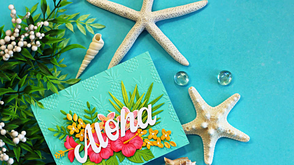 Handmade tropical Aloha card featuring layered hibiscus flowers, tropical leaves, and a large white “Aloha” sentiment on an aqua embossed background. The card is styled with starfish, seashells, greenery, and glass gems on a bright turquoise backdrop, creating a relaxing island-inspired scene.