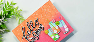 Bright summer card featuring a pink and orange ink-blended background with a layered pool-water stencil design. The card is decorated with colorful die cut summer drinks, citrus slice embellishments, and a bold black and white “hello sunshine” sentiment for a fun tropical vibe.
