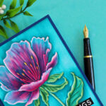Close-up of a handmade floral greeting card featuring the Simon Says Stamp Curvy Blossom Embossing Folder. The embossed flower is colored with Copic markers on the debossed side, with blue ink dragged over the raised surface to highlight the texture. The card is displayed on a turquoise background with greenery and a fountain pen.