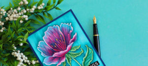 Close-up of a handmade floral greeting card featuring the Simon Says Stamp Curvy Blossom Embossing Folder. The embossed flower is colored with Copic markers on the debossed side, with blue ink dragged over the raised surface to highlight the texture. The card is displayed on a turquoise background with greenery and a fountain pen.
