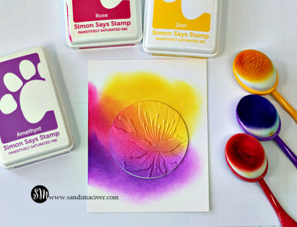 White cardstock panel featuring the Spotlight Technique with Embossing Folders using the Simon Says Stamp Curvy Blossom 3D Embossing Folder. Bright ink blending in yellow, pink, and purple surrounds a circular embossed flower spotlight, with Simon Says Stamp ink pads and blending brushes shown alongside.