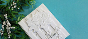 White-on-white embossed floral card featuring the Embossed Double Vision technique. A large dimensional flower and delicate floral details are layered over raised vertical panels, creating elegant depth and texture against a soft turquoise background.