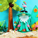 Handmade tropical felt décor featuring Fergus the gnome decorated with pink flamingos and hibiscus flowers. Fergus stands on a sandy beach beside a felt palm tree and surfboard, surrounded by seashells and greenery for a colorful summer paradise scene.