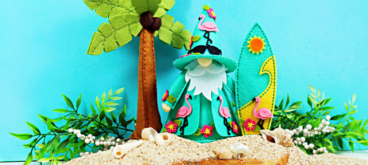 Handmade tropical felt décor featuring Fergus the gnome decorated with pink flamingos and hibiscus flowers. Fergus stands on a sandy beach beside a felt palm tree and surfboard, surrounded by seashells and greenery for a colorful summer paradise scene.