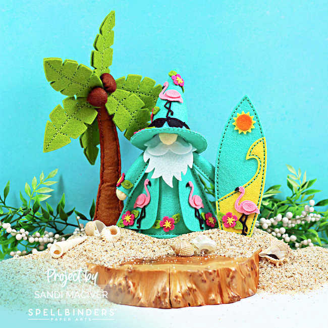 Tropical felt gnome décor featuring Fergus dressed in teal and decorated with pink flamingos and hibiscus flowers. The handmade summer display includes a stitched palm tree, surfboard, sandy beach, seashells, and lush greenery for a colorful island-inspired scene.