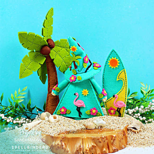 Back view of Fergus the tropical felt gnome decorated with pink flamingos, hibiscus flowers, sunshine accents, and tropical foliage. The handmade summer décor scene features a stitched palm tree, surfboard, sandy beach, and seashells, showcasing the detailed felt embellishments from every angle.