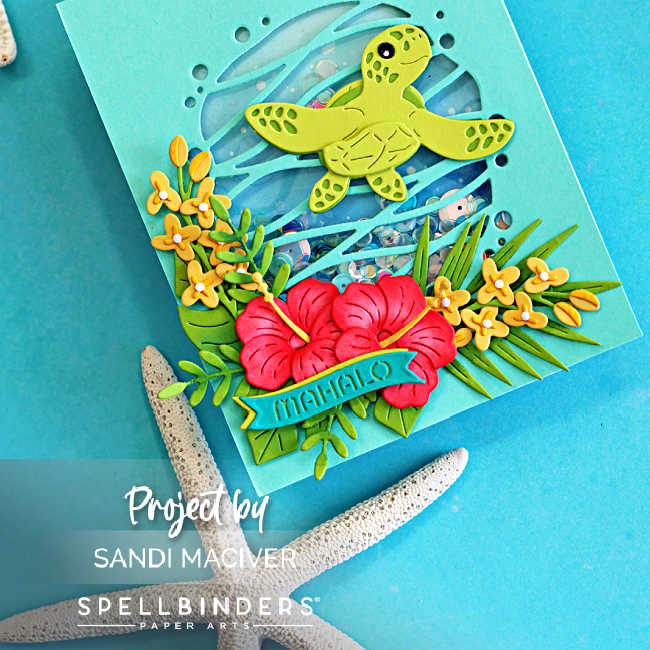 Close-up of a tropical handmade shaker card featuring a layered green sea turtle swimming across a blue ocean-inspired shaker window filled with sequins. Bright pink hibiscus flowers, yellow tropical blossoms, and lush green foliage cluster along the bottom of the card, with a layered Mahalo sentiment nestled among the flowers. The card is photographed on a turquoise background with a starfish accent, creating a vibrant Hawaiian paradise theme.