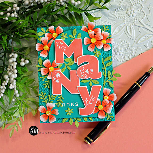 Handmade floral thank you card featuring oversized coral "Many" die-cut letters layered over a teal arched panel with a subtle embossed background. Coral flowers with yellow centers and green foliage frame the sentiment, with a black fountain pen beside the card.