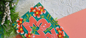 Close-up of a layered handmade thank you card featuring oversized coral "Many" letters surrounded by coral flowers and green foliage on a teal embossed arched background. The card is accented with a textured white embossed backdrop and soft peach cardstock.