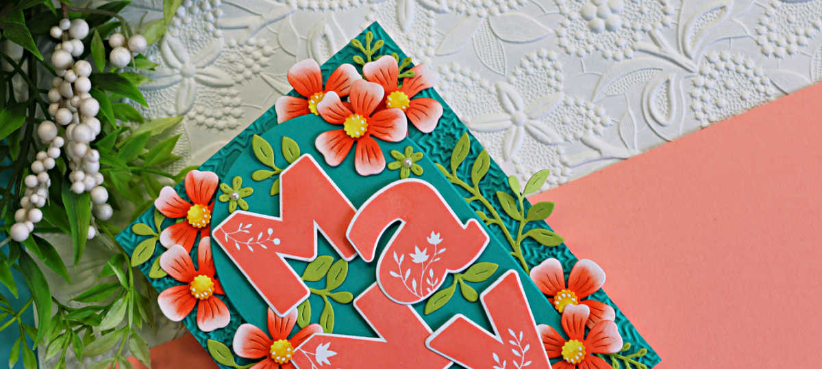 Close-up of a layered handmade thank you card featuring oversized coral "Many" letters surrounded by coral flowers and green foliage on a teal embossed arched background. The card is accented with a textured white embossed backdrop and soft peach cardstock.