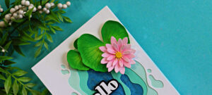 Close-up of a handmade layered pond lily card featuring dimensional green lily pads and a pink water lily flower over a multi-layered aqua and blue pond background. The card is photographed at an angle against a soft turquoise backdrop with leafy greenery in the upper left corner.