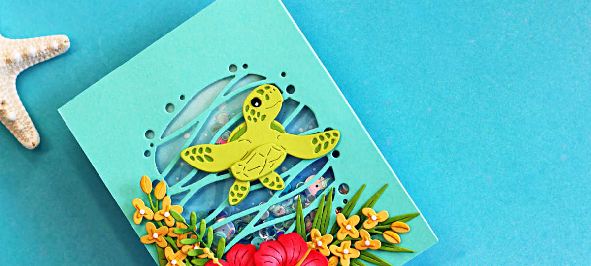 Close-up of a tropical handmade shaker card featuring a layered sea turtle swimming over a blue ocean shaker window filled with sequins. Bright hibiscus flowers, tropical foliage, and yellow blossoms frame the bottom of the design, creating a colorful Hawaiian paradise theme on a soft aqua card background.