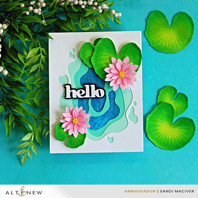 Handmade layered pond lily card featuring a dimensional blue and aqua pond scene created with stacked die-cut layers. Two pink water lilies with yellow centers rest among green lily pads, with a bold black-and-white "hello" sentiment across the center. The card is displayed on a turquoise background surrounded by additional lily pads and leafy greenery.