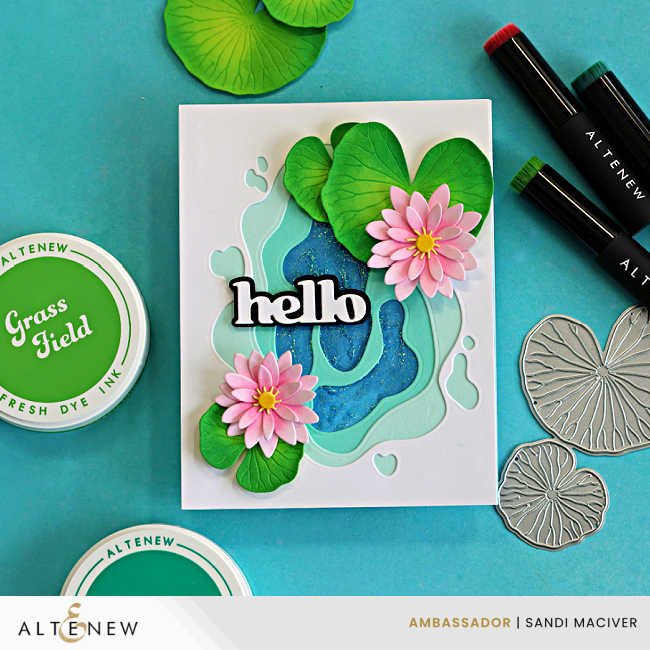 Handmade layered pond lily card featuring a dimensional aqua and blue pond created with stacked die-cut layers. Two pink water lilies with yellow centers sit among green lily pads, with a bold black-and-white "hello" sentiment across the center. The card is displayed with Altenew ink pads, blending brushes, and coordinating lily pad dies on a turquoise background.