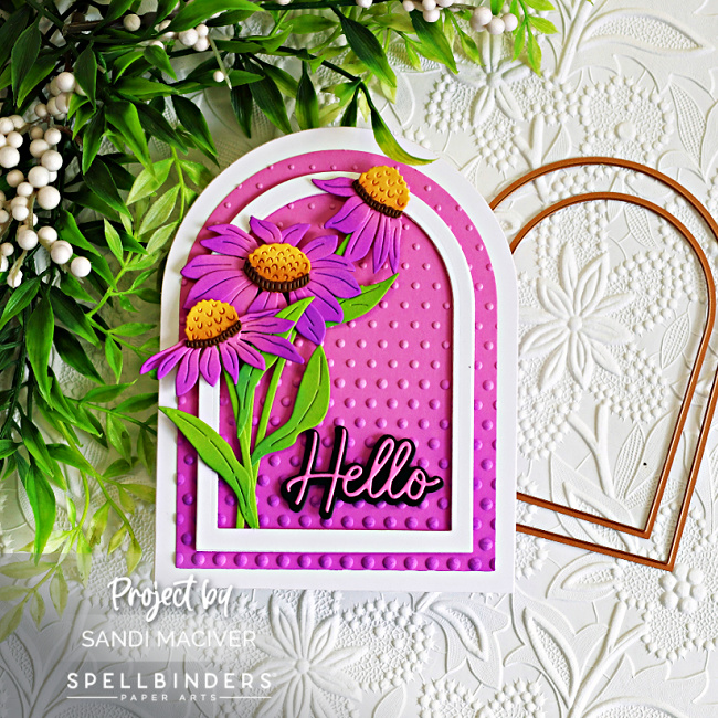 Handmade arched greeting card featuring layered purple wild cornflowers with bright green leaves and golden-yellow flower centers. The flowers are arranged inside a white arched frame over a vibrant pink embossed background created with the Layered Cascading Dots Embossing Folder. A bold black "Hello" sentiment anchors the design, while a white embossed floral background, greenery, and a black fountain pen add an elegant garden-inspired feel. 🌸💗💜