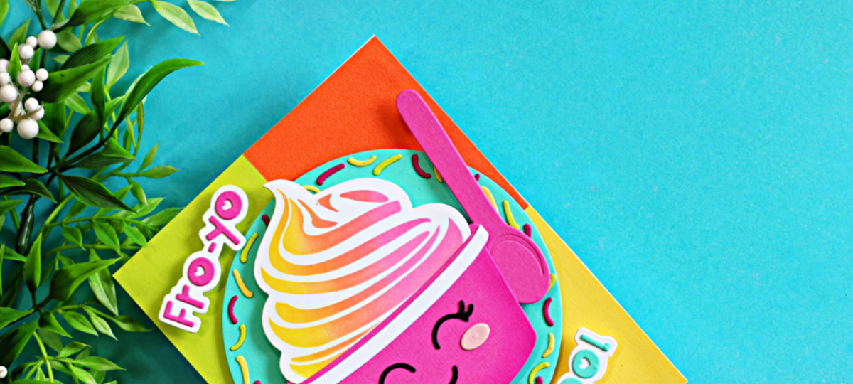 Close-up of a colorful handmade card featuring a smiling frozen yogurt cup die cut from the Trinity Stamps Fro-Yo Friends release. The fro-yo is stenciled in soft pink, orange, and yellow hues, accented with a bright pink spoon and playful sprinkle frame. The card sits on a vibrant color-blocked background of pink, yellow, orange, and aqua cardstock, creating a fun and cheerful summer design.