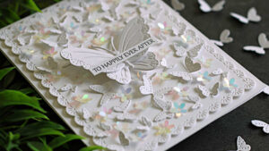 Elegant all-white butterfly wedding card featuring layered die-cut butterflies, iridescent floral embellishments, and a delicate scalloped frame. A dimensional butterfly centerpiece carries a "To Happily Ever After" sentiment, while the card is photographed on a dark background with scattered butterflies and greenery.