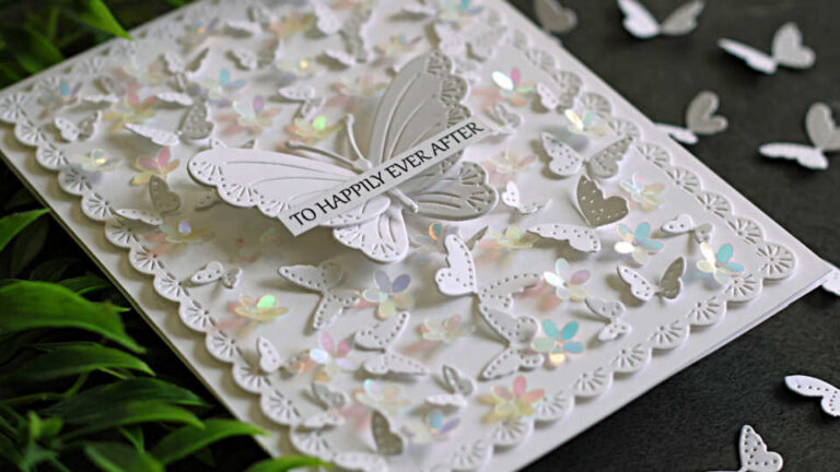 Elegant all-white butterfly wedding card featuring layered die-cut butterflies, iridescent floral embellishments, and a delicate scalloped frame. A dimensional butterfly centerpiece carries a "To Happily Ever After" sentiment, while the card is photographed on a dark background with scattered butterflies and greenery.