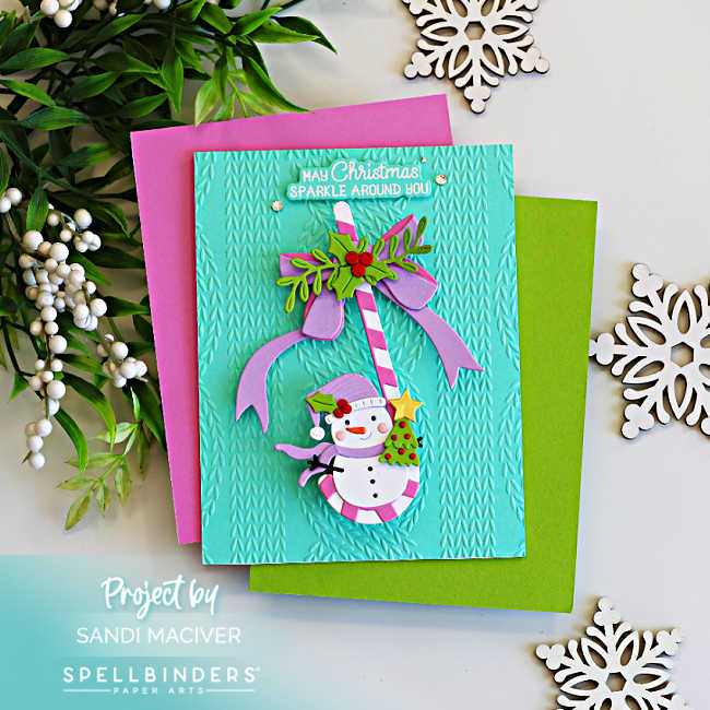 Interactive Christmas card featuring a swinging snowman hanging from a pink and white candy cane on a turquoise knit embossed background, layered over bright pink and green cardstock with festive snowflake accents.