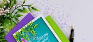 Close-up of a handmade floral shaker card featuring the Simon Says Stamp Leaf Dance Flowers and Frame die. Ink-blended green foliage and purple flowers surround a teal shaker window filled with clear sequins, with a white embossed sentiment reading "Celebrating another year to bloom." The card is styled with purple and green cardstock, a black fountain pen, scattered iridescent gems, and greenery on a white background.
