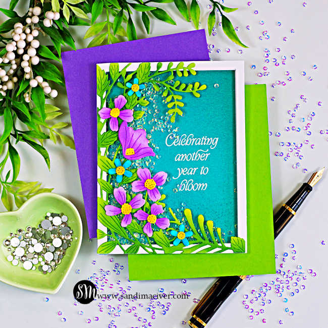 Handmade easy shaker card featuring the Simon Says Stamp Leaf Dance Flowers and Frame die. Ink-blended green foliage and purple flowers frame a teal shaker window filled with sparkling clear sequins. The white embossed sentiment reads, "Celebrating another year to bloom." The card is photographed on purple and lime green cardstock with a fountain pen, heart-shaped dish of gems, scattered iridescent embellishments, and greenery on a white background.