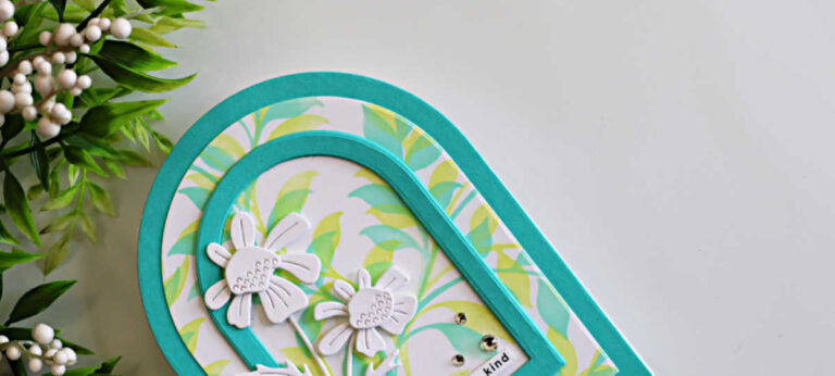 Close-up of a handmade arched greeting card featuring a soft pastel leafy green stenciled background framed with teal A2 arches. White layered die-cut flowers with embossed details decorate the lower left corner, accented with a few gold gems for sparkle.