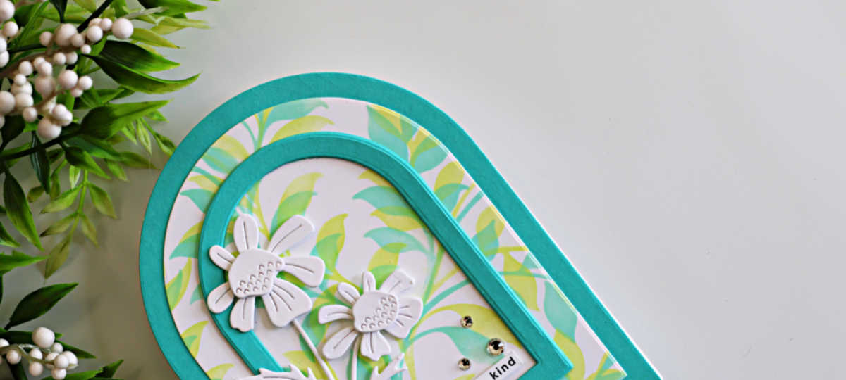 Close-up of a handmade arched greeting card featuring a soft pastel leafy green stenciled background framed with teal A2 arches. White layered die-cut flowers with embossed details decorate the lower left corner, accented with a few gold gems for sparkle.