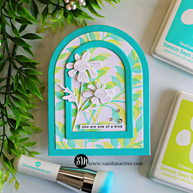 Handmade arched greeting card featuring a soft pastel Leafy Greens stenciled background, layered teal A2 arch frames, and white die-cut daisy flowers with embossed details. The card is displayed beside the Simon Says Stamp Quirky Daisies die set and a blending brush, with a sentiment reading, "you are one of a kind."