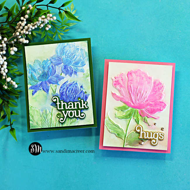 Two handmade faux watercolor floral cards created with embossed backgrounds and Karin Brushmarker Pro markers. One card features blue flowers with a layered "thank you" die-cut sentiment on a dark green card base, while the second showcases a pink flower with a layered "hugs" sentiment on a bright pink card base. Both cards have soft watercolor shading, subtle paint splatter, and sparkling gems.