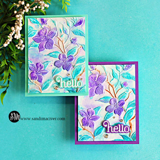 Two handmade faux watercolor floral cards created with embossed backgrounds and Karin Brushmarker Pro markers. The cards feature purple flowers with aqua leaves, soft watercolor shading, and layered "hello" die-cut sentiments on teal and purple card bases. Finished with subtle paint splatter and sparkling gems, the cards are displayed on a turquoise background with greenery in the upper left corner.