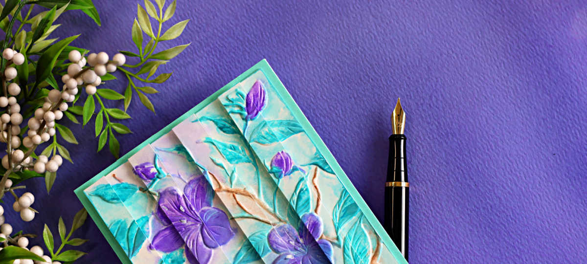 Close-up of a handmade floral card featuring the Faux Watercolor Embossed Double Vision technique with purple flowers, aqua embossed leaves, and a turquoise card base on a purple background.