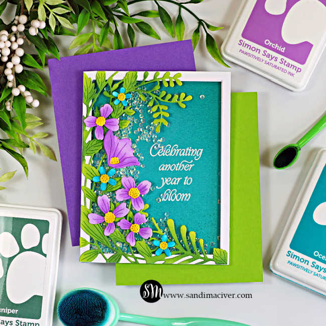 Handmade easy shaker birthday card featuring the Simon Says Stamp Leaf Dance Flowers and Frame die. Ink-blended green foliage and purple flowers surround a teal shaker window filled with sparkling clear sequins. The white embossed sentiment reads, "Celebrating another year to bloom." The card is styled with Simon Says Stamp ink pads, blending brushes, and colorful cardstock on a white background.
