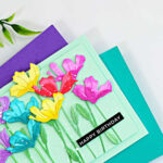 Colorful handmade birthday card featuring rainbow die-cut flowers created with the Simon Says Stamp Tissue Petal Stems die. The ink-blended flowers are arranged on a soft mint green background with layered stems, white paint splatter, and a black "Happy Birthday" sentiment. The card is displayed on teal and purple cardstock with leafy greenery in the corner.