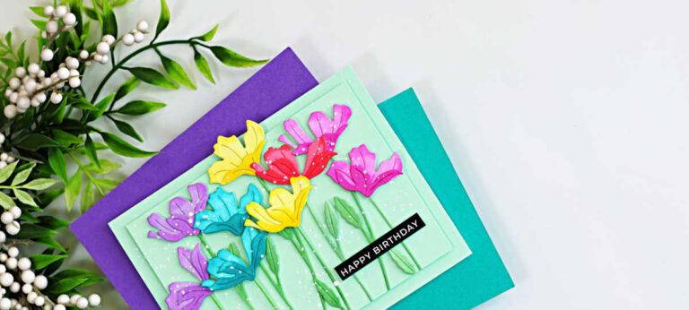 Colorful handmade birthday card featuring rainbow die-cut flowers created with the Simon Says Stamp Tissue Petal Stems die. The ink-blended flowers are arranged on a soft mint green background with layered stems, white paint splatter, and a black "Happy Birthday" sentiment. The card is displayed on teal and purple cardstock with leafy greenery in the corner.