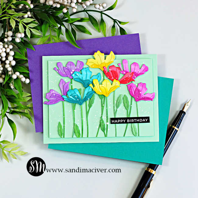 Handmade birthday card featuring rainbow die-cut flowers created with the Simon Says Stamp Tissue Petal Stems die. Ink-blended flowers in purple, teal, pink, red, and yellow bloom against a soft mint green background with layered green stems, white paint splatter, and a black "Happy Birthday" sentiment. The card is displayed on teal and purple cardstock beside a black fountain pen and leafy greenery.