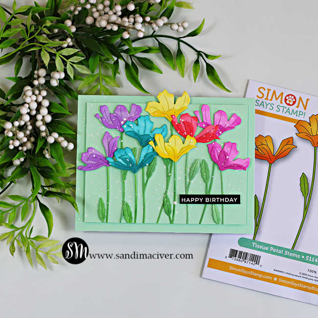 Handmade rainbow die-cut flower birthday card created with the Simon Says Stamp Tissue Petal Stems die. Ink-blended flowers in purple, teal, yellow, red, and pink are arranged on a soft mint green background with layered green stems, white paint splatter, and a black "Happy Birthday" sentiment. The coordinating Tissue Petal Stems die package is displayed beside the card.