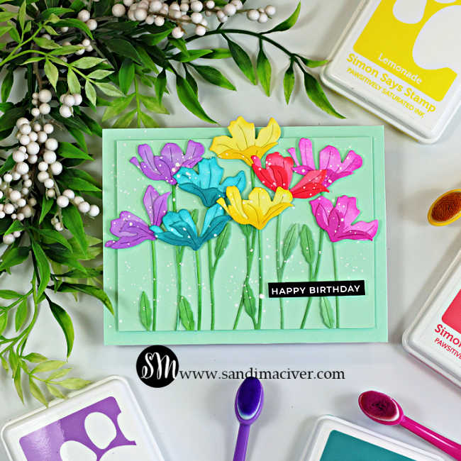Handmade rainbow die-cut flower birthday card featuring ink-blended flowers created with the Simon Says Stamp Tissue Petal Stems die. Purple, teal, yellow, red, and pink flowers bloom on a soft mint green background with layered stems, white paint splatter, and a black "Happy Birthday" sentiment. Simon Says Stamp ink pads and blending brushes surround the card.