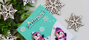 Handmade Christmas card featuring two adorable die-cut penguins in purple hats and scarves playing on snowy hills beneath a teal sky. White snowflakes, splattered snow, and the "Let It Snow" sentiment complete this festive card created with the Spellbinders Snow Much Fun Small Die of the Month.