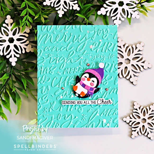 Handmade Christmas card featuring a cute die-cut penguin wearing a purple hat and scarf on a teal background embossed with the Spellbinders Festive Greetings 3D Embossing Folder. The card is finished with a "Sending You All the Cheer" sentiment and sparkling gems.