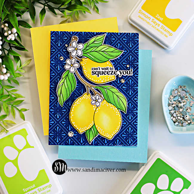 A handmade greeting card featuring two bright yellow lemons with green leaves and white blossoms, layered over a deep navy blue embossed background with a white-highlighted tile pattern. The sentiment, "can't wait to squeeze you!", is nestled across the top lemon. The card is displayed on layered yellow and light blue cardstock and surrounded by coordinating Simon Says Stamp ink pads, a dish of clear gems, and greenery on a white work surface.