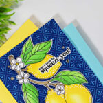Close-up of a handmade lemon-themed greeting card resting on layers of yellow and light blue cardstock. The card features bright yellow lemons, green leaves, and white blossoms on a deep navy embossed background highlighted with white pigment ink to resemble Spanish ceramic tiles. The sentiment, "can't wait to squeeze you!", is placed across the top lemon. Decorative greenery frames the upper left corner against a clean white background.