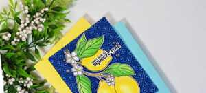 Close-up of a handmade lemon-themed greeting card resting on layers of yellow and light blue cardstock. The card features bright yellow lemons, green leaves, and white blossoms on a deep navy embossed background highlighted with white pigment ink to resemble Spanish ceramic tiles. The sentiment, "can't wait to squeeze you!", is placed across the top lemon. Decorative greenery frames the upper left corner against a clean white background.