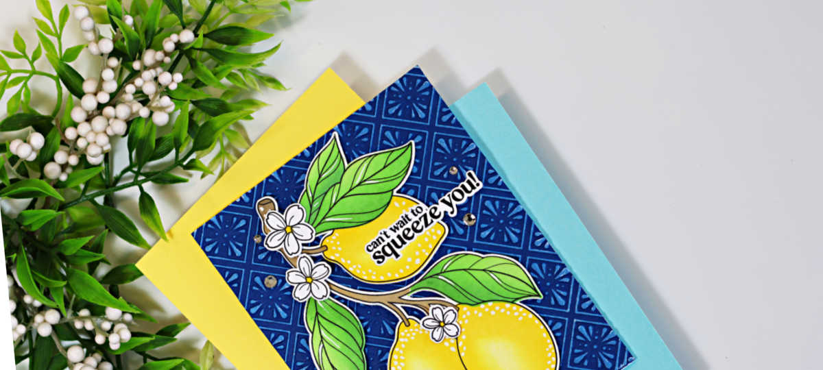 Close-up of a handmade lemon-themed greeting card resting on layers of yellow and light blue cardstock. The card features bright yellow lemons, green leaves, and white blossoms on a deep navy embossed background highlighted with white pigment ink to resemble Spanish ceramic tiles. The sentiment, "can't wait to squeeze you!", is placed across the top lemon. Decorative greenery frames the upper left corner against a clean white background.