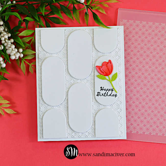 White birthday card featuring a geometric oval die-cut design over a textured Woven Rings embossed background, accented with a coral flower and shown beside the embossing folder.