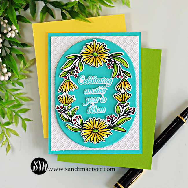 A handmade birthday card featuring the Simon Says Stamp Daisy Frame, colored with bright yellow flowers and green foliage using Copic markers. The floral frame surrounds the sentiment, "Celebrating another year to bloom," on an Audrey Blue oval die cut with the Chunky Ovals. The focal panel is layered over a white background embossed with the Woven Rings Embossing Folder, with yellow and green cardstock and a black pen arranged beneath the card.