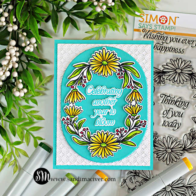 A handmade birthday card featuring the Simon Says Stamp Daisy Frame, colored with bright yellow and green Copic markers and die cut with the Chunky Ovals. The floral frame surrounds the sentiment, "Celebrating another year to bloom," stamped in Surf Ink on an Audrey Blue oval. The card is layered over a white panel embossed with the Woven Rings Embossing Folder and displayed alongside the Daisy Frame stamp set and Copic markers.