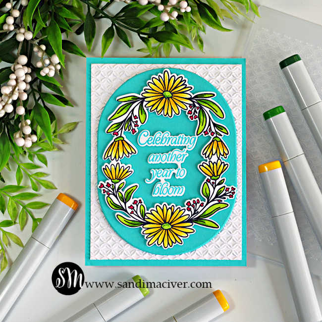 A handmade birthday card featuring the Simon Says Stamp Daisy Frame, colored with bright yellow and green Copic markers. The floral wreath frames the sentiment, "Celebrating another year to bloom," on an Audrey Blue oval die cut with the Chunky Ovals. The focal panel is layered over a white background embossed with the Woven Rings Embossing Folder, with Copic markers and greenery arranged around the card for a clean crafting flat lay.